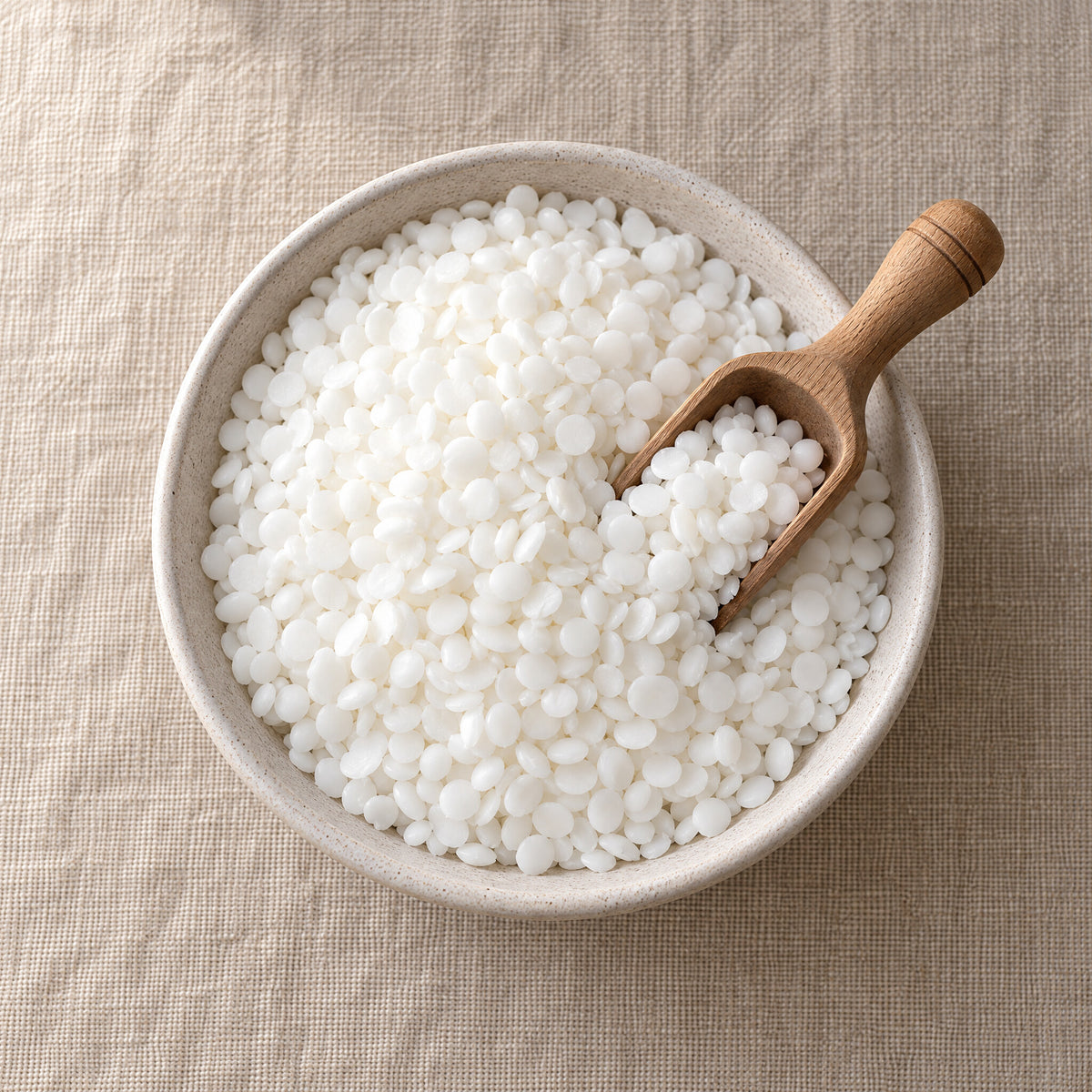 Soy pillar wax beads in a shallow bowl with a wooden scoop, seen from above on a linen surface