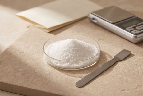 A glass dish of white ambroxan crystals beside a steel spatula and a pocket scale on a stone worktop