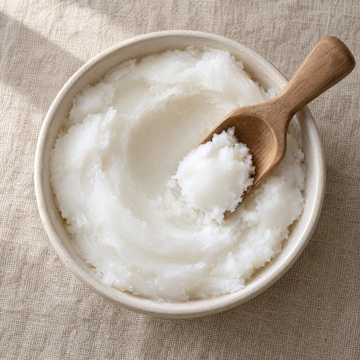 Coconut wax in a shallow bowl with a wooden scoop, seen from above on a linen surface