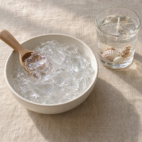 Clear gel wax in a shallow bowl with a wooden scoop, seen from above on a linen surface
