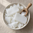 Fully refined paraffin wax pieces in a shallow bowl with a wooden scoop, seen from above on a linen surface